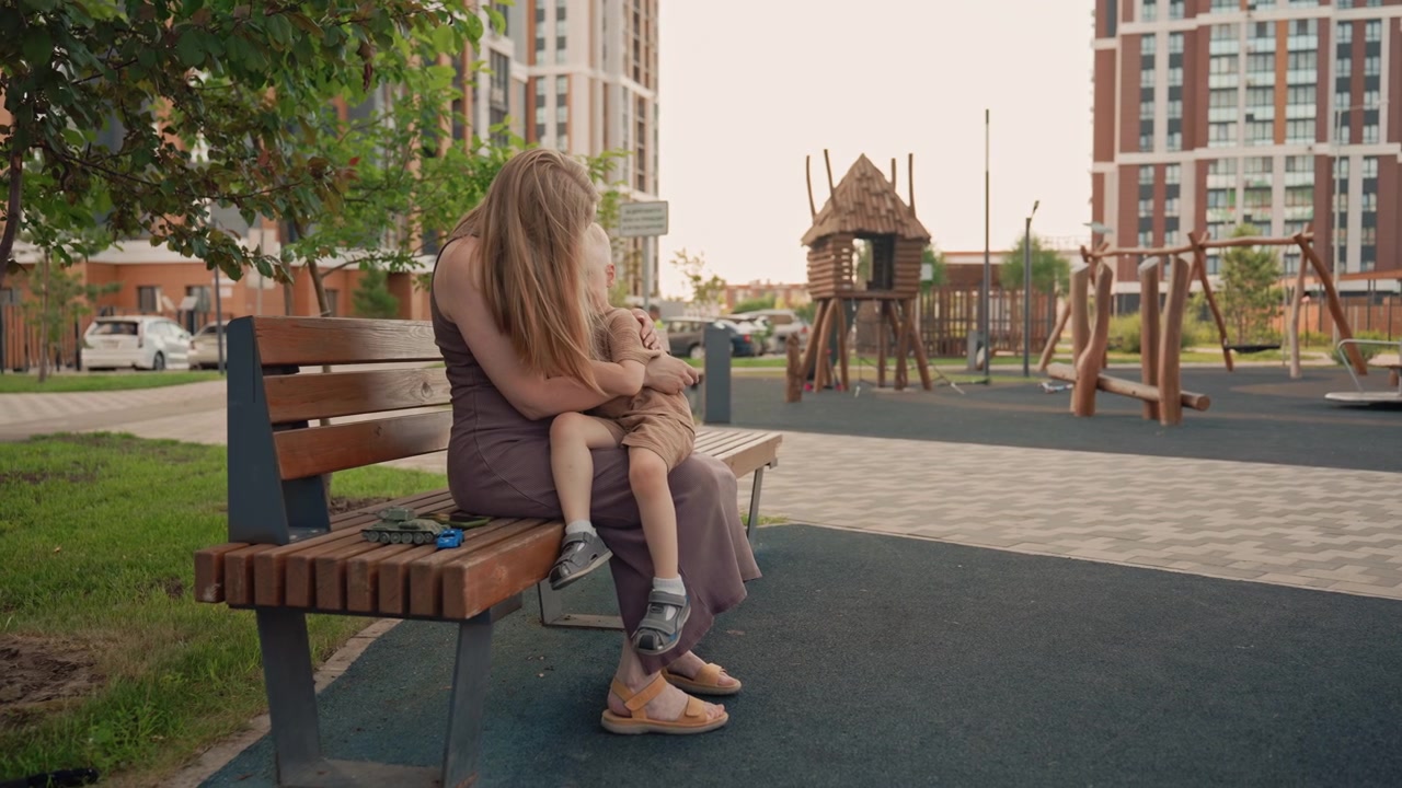 Donna seduta su una panchina in un parco urbano che tiene e conforta un bambino piccolo. La scena include veicoli giocattolo posizionati accanto a loro con attrezzature da parco giochi ed edifici residenziali visibili sullo sfondo
