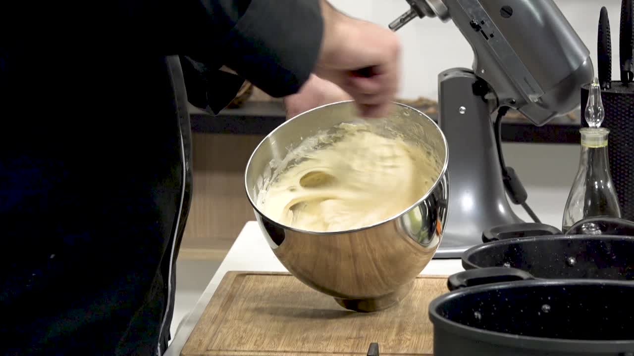 A chef prepares a delicious cake batter, using a stainless steel bowl. Mixing ingredients thoroughly, the kitchen is filled with the aroma of baking. A relaxed atmosphere prevails.