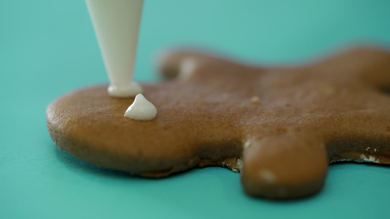Confectioner is painting gingerbread boy with white glaze. Decor by icing of delicious gingerbread in the form of a boy with eyes, mouth on the blue background. Close-up