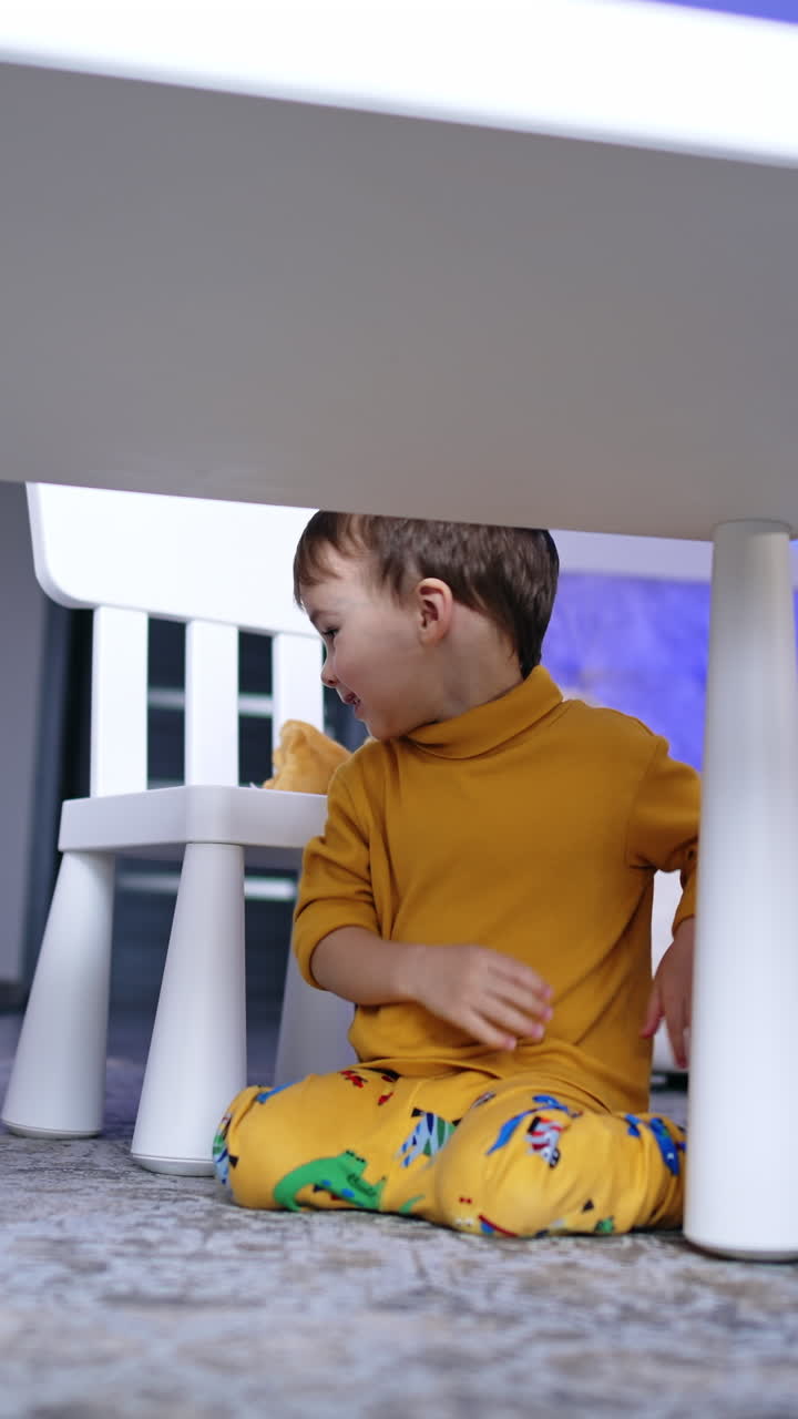 Bambino di due anni seduto sulle ginocchia sul pavimento vicino alla scrivania. Neonati in abiti gialli gattonano sul pavimento. Video verticale