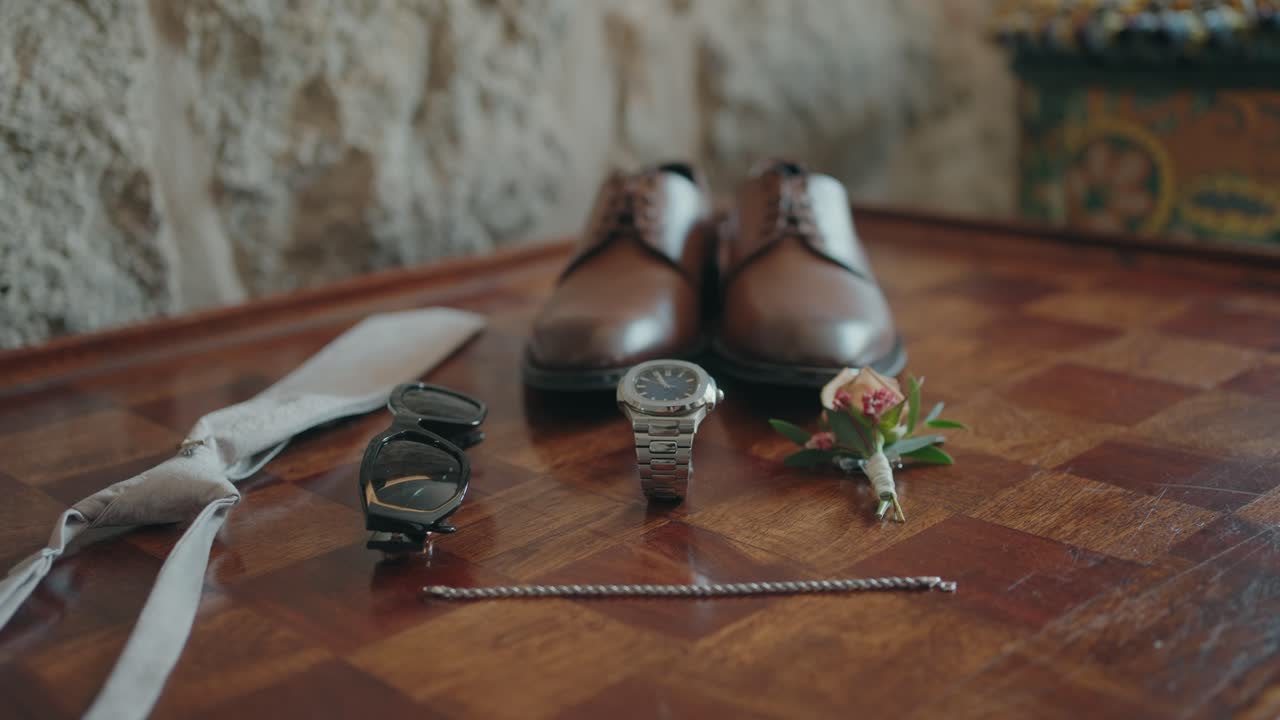 Elegant flat lay of groom’s shoes, watch, tie, sunglasses, bracelet and boutonniere