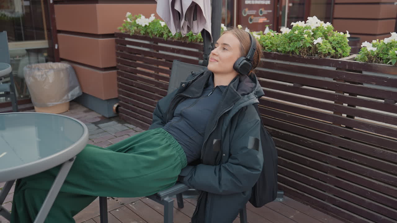 Ung, hvid kvinde slapper af på en udendørs café med hovedtelefoner, grønt skirt og mørk jakke. Hun læner sig tilbage i stolen ved siden af et metalbord og plantekasser, med et roligt smil og et blidt blik. Freelanceskribent holder en