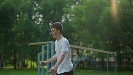 a young boy in a white shirt, intensely focused on his outdoor training as he jumps, with various sports equipment and trees in the background