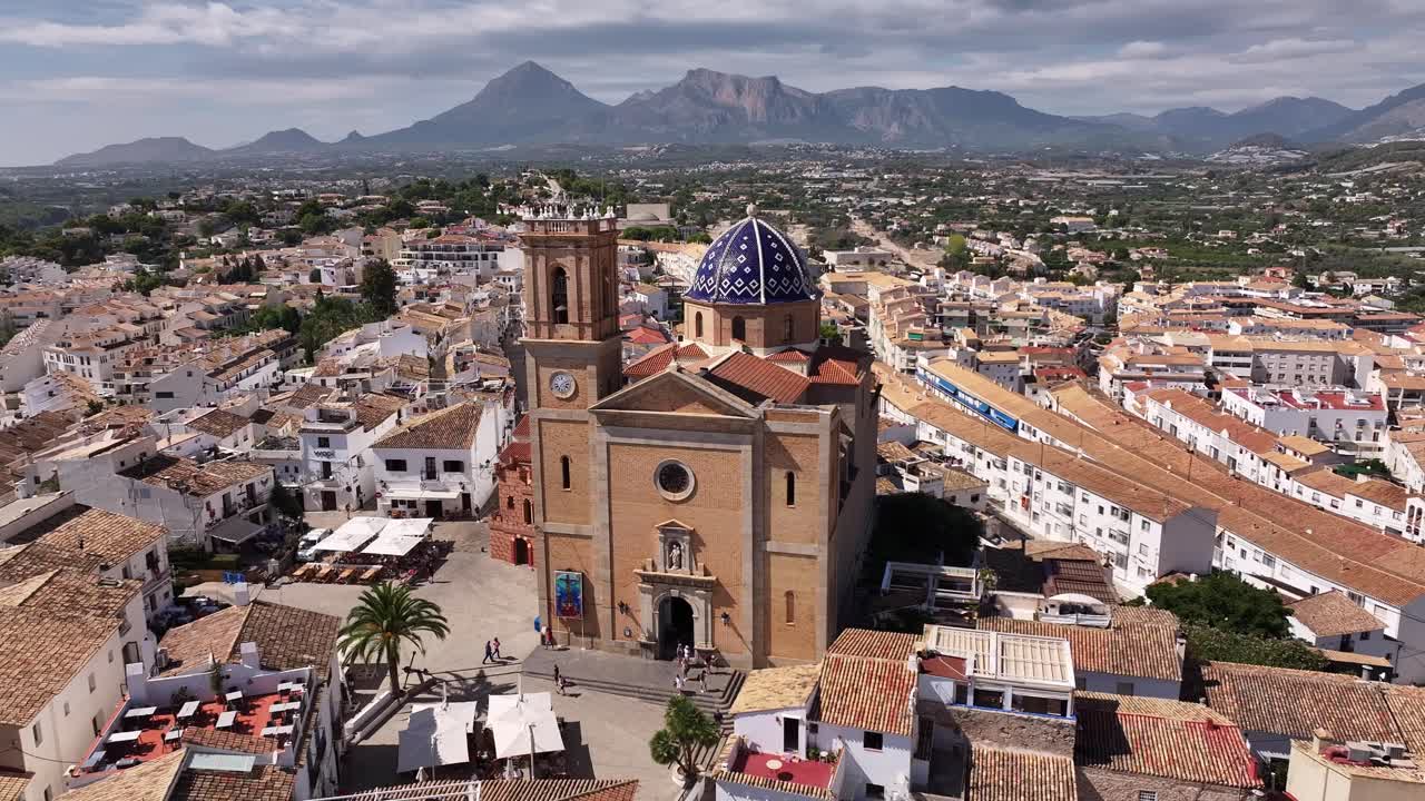 Aerial orbiting view over Altea captures the blue-domed church of Nuestra Señora del Consuelo with the Sierra de Bernia mountain ridge in the background, tiled roofs below, real time, drone orbit