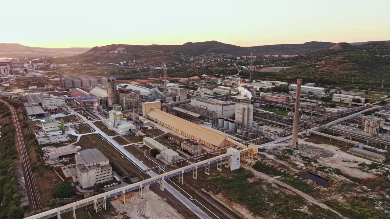 Complexo industrial localizado em uma área montanhosa perto de uma cidade ao pôr do sol com fumaça saindo de chaminés e vários edifícios presentes