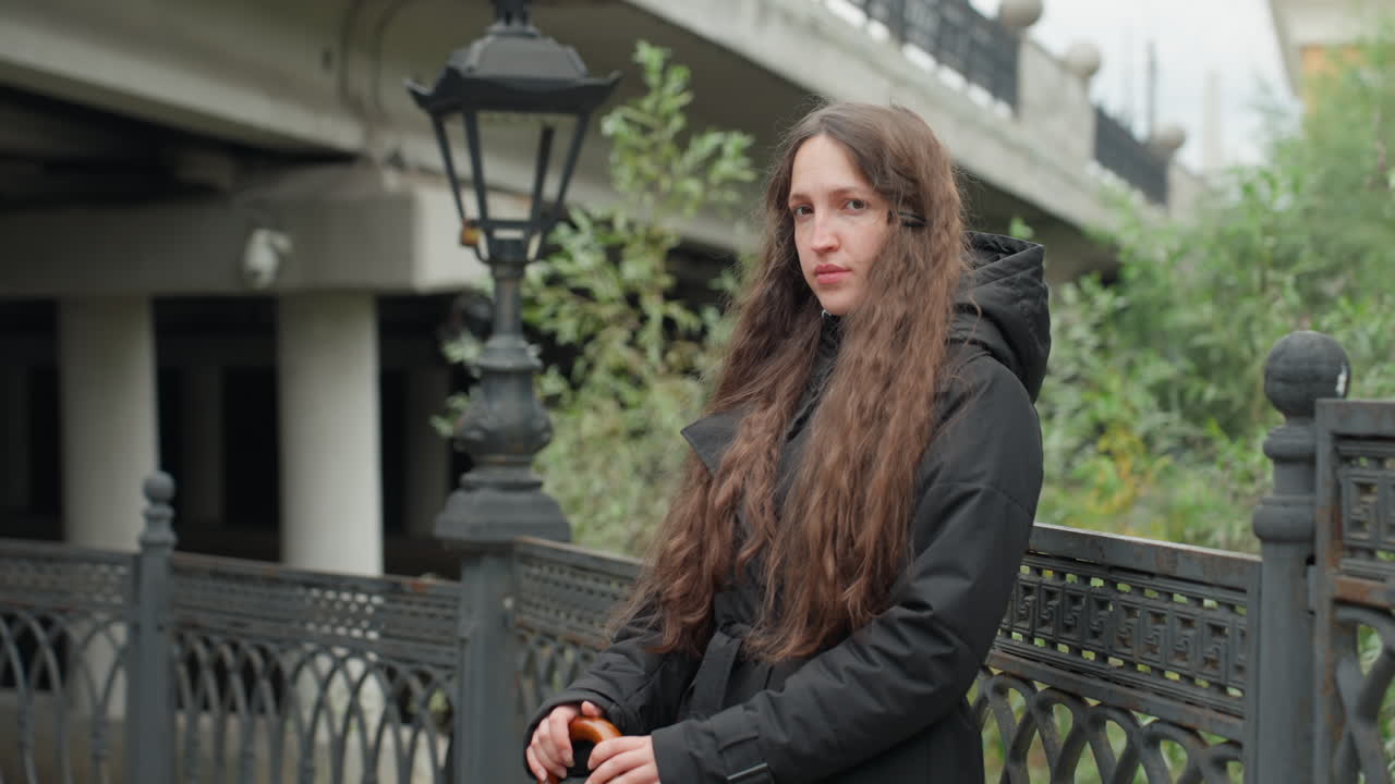 Donna bianca a figura intera appoggiata a una ringhiera, in piedi sotto un cavalcavia con lunghi capelli castani e un cappotto nero, che tiene in mano un piccolo oggetto vicino a una recinzione di ferro e un lampione vintage, pausa pendolare urbana con sguardo contemplativo