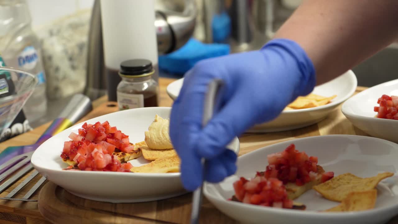 el chef con guantes de goma azules coloca hummus en chips de pan de pita con tomates cortados en cubitos, armando un aperitivo saludable en la cocina usando una cuchara y tazones o platos blancos, comida cerrada