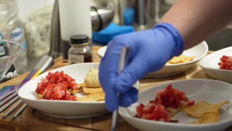 Chef-Wearing-Blue-Rubber-Gloves-Places-Hummus-on-Pita-Bread-Chips-with-Diced-Tomatoes,-Assembling-a-Healthy-Appetizer-Meal-by-in-the-Kitchen-using-a-Spoon-and-White-Bowls-or-Plates,-Food-Closeup
