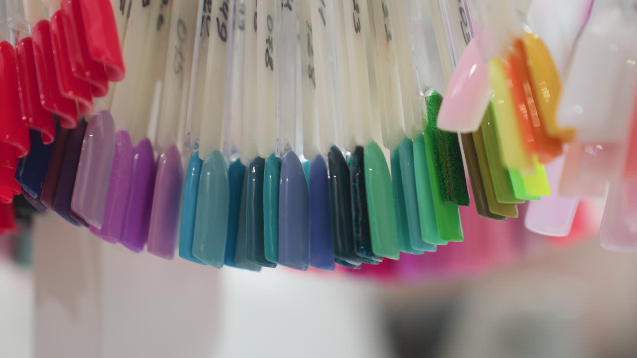 Close up view of colorful nail polish swatches hanging on plastic sticks with visible handwritten labels, featuring vibrant shades and gradients in soft blur background inside beauty salon