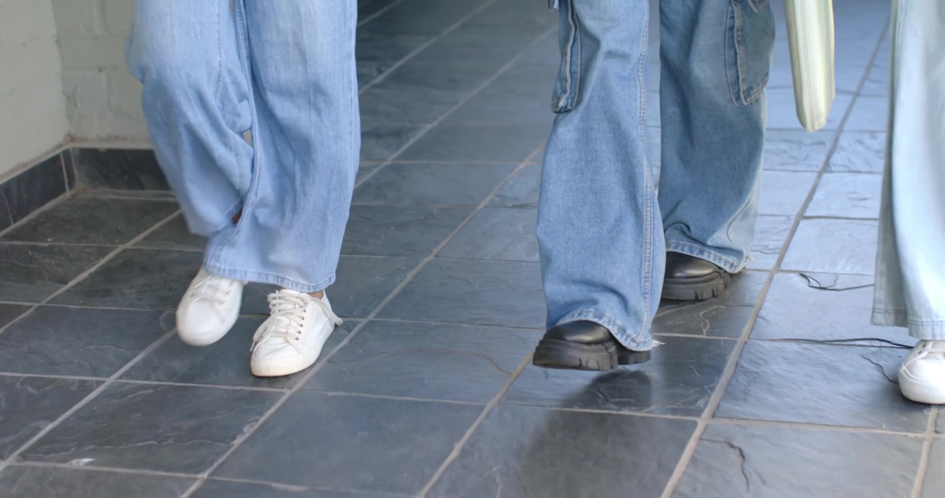 Due donne e un bambino che indossano pantaloni di jeans avanzano lungo un corridoio piastrellato, portando una borsa verde chiaro.