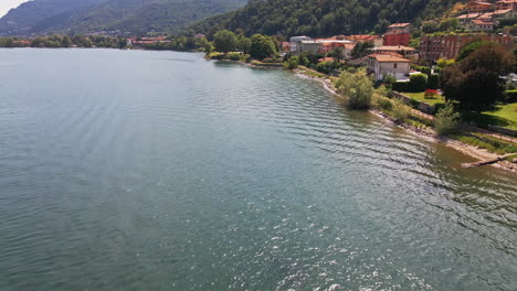 calm waters of lake como with a view of the shore and residential houses with green mountains in background at daytime in italy - aerial drone forward