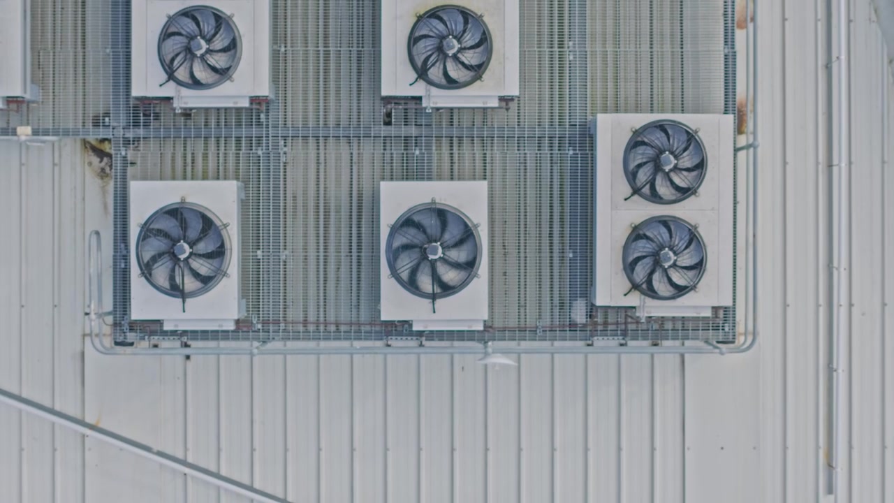 Dronevideo toont een breed shot van meerdere grote koelventilatoren en HVAC-units op het dak van een modern datacenter. Schone industriële omgeving, metallic tinten, geometrische patronen en statische compositie