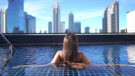 Rückseite-Einer-Stilvollen-Frau-Mit-Langen-Haaren,-Die-Sich-Im-Pool-Auf-Dem-Dach-Mit-Atemberaubendem-Blick-Auf-Die-Moderne-Stadt-Und-Die-Wolkenkratzer-Von-Bangkok,-Vollbild,-Genießt