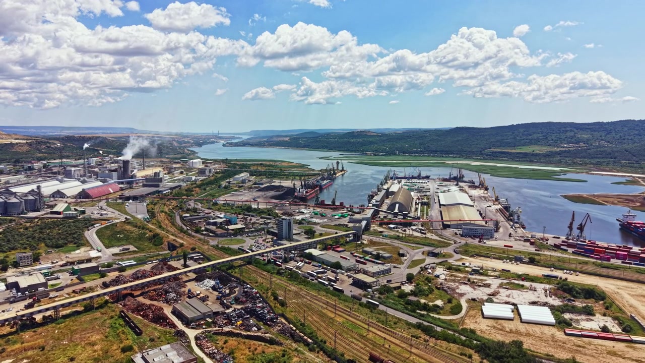 Vista de una zona industrial junto a un río con buques de carga, fábricas y una autopista en un paisaje rural bajo un cielo nublado.