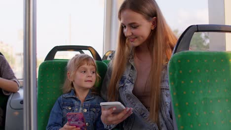 Mother-with-daughter-traveling-by-bus-going-home,-playing-on-smartphone,-browsing-social-media