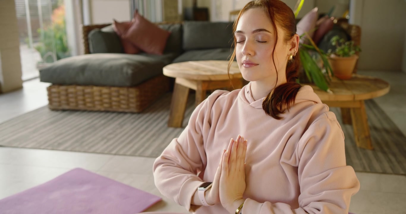 Donna con felpa rosa che inspira, alza le braccia, le incrocia al petto, meditando su un tappetino da yoga a casa.