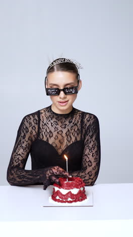 birthday girl wearing black long-sleeve bodysuit and diadem sits at the table looking at cake. woman is given sunglasses and a lit candle which she puts into cake and blows. vertical video.