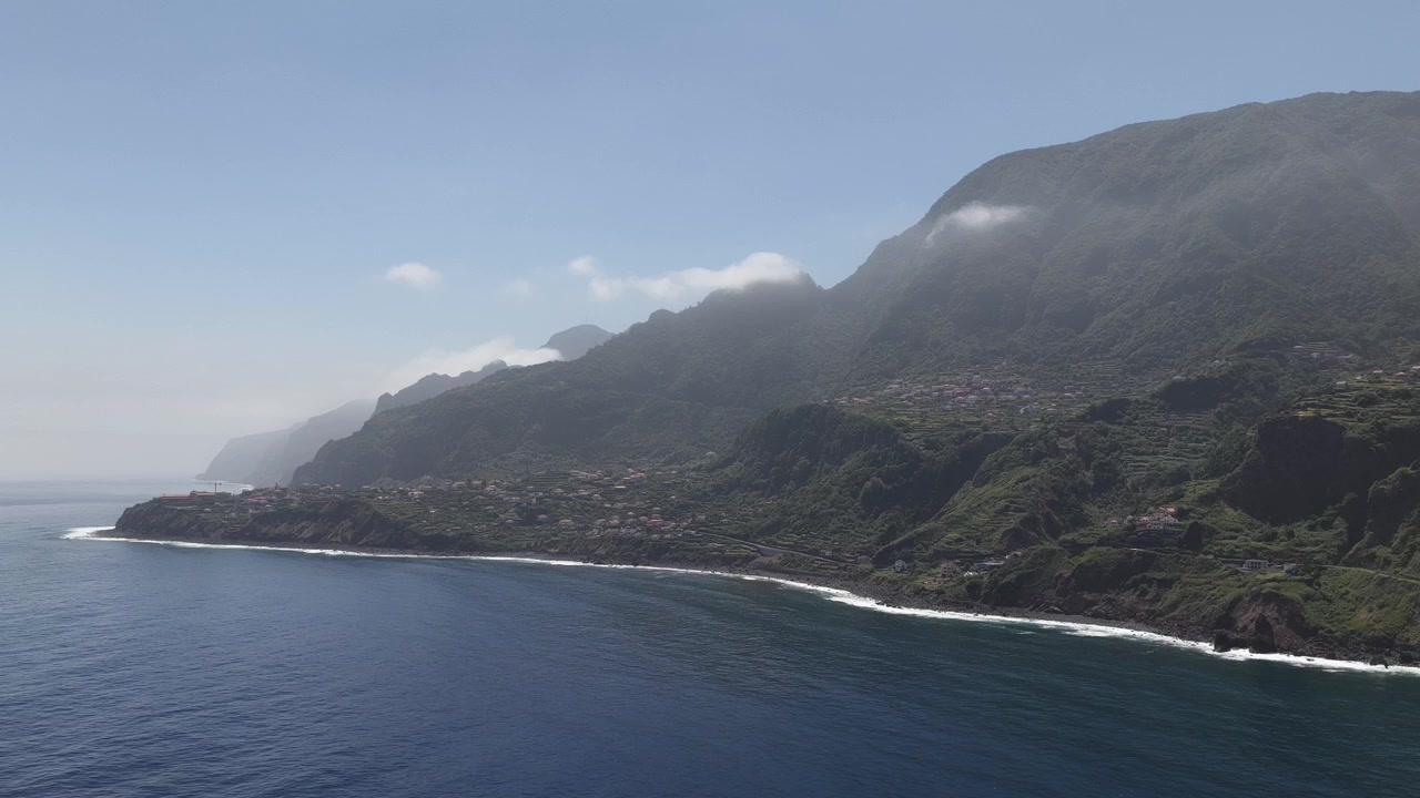 Una vista aerea cattura le lussureggianti scogliere e la costa vulcanica dell'isola di Madeira, Portogallo, con la nebbia mattutina che si estende su ripide montagne verdi e il profondo blu dell'Atlantico alla luce nitida del giorno.