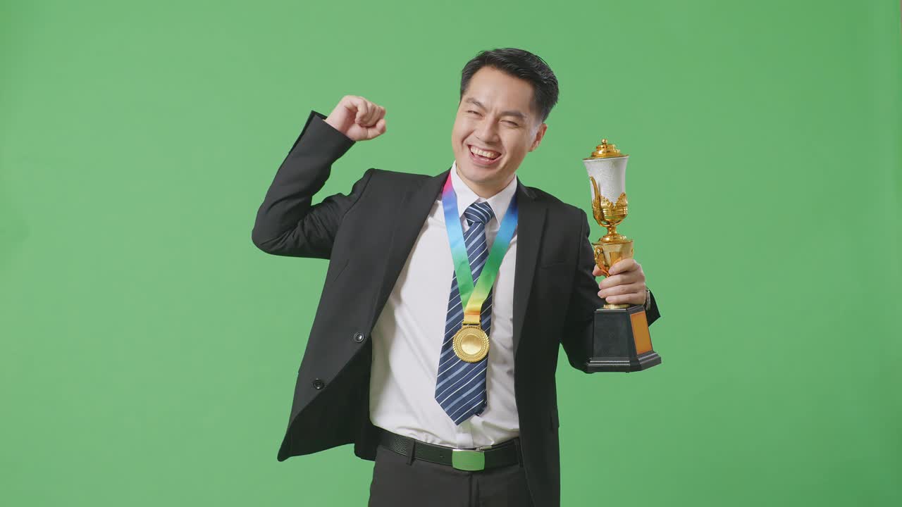 슈트와 타이를 입은 아시아 사업가, 금메달과 트로피를 들고, 양팔을 구부리고 스튜디오의 녹색 스크린 배경에서 카메라에 미소 짓고