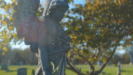 A-beautiful-shot-of-a-tombstone-adorned-with-a-concrete-angel-statue-holding-a-plastic-flower,-against-tombs-in-the-back-and-under-a-blue-sky