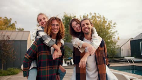 Portrait:-two-girls-are-sitting-on-the-backs-of-their-boyfriends-in-checkered-shirts-and-smiling-near-sunbeds.-Rest-in-the-country-house