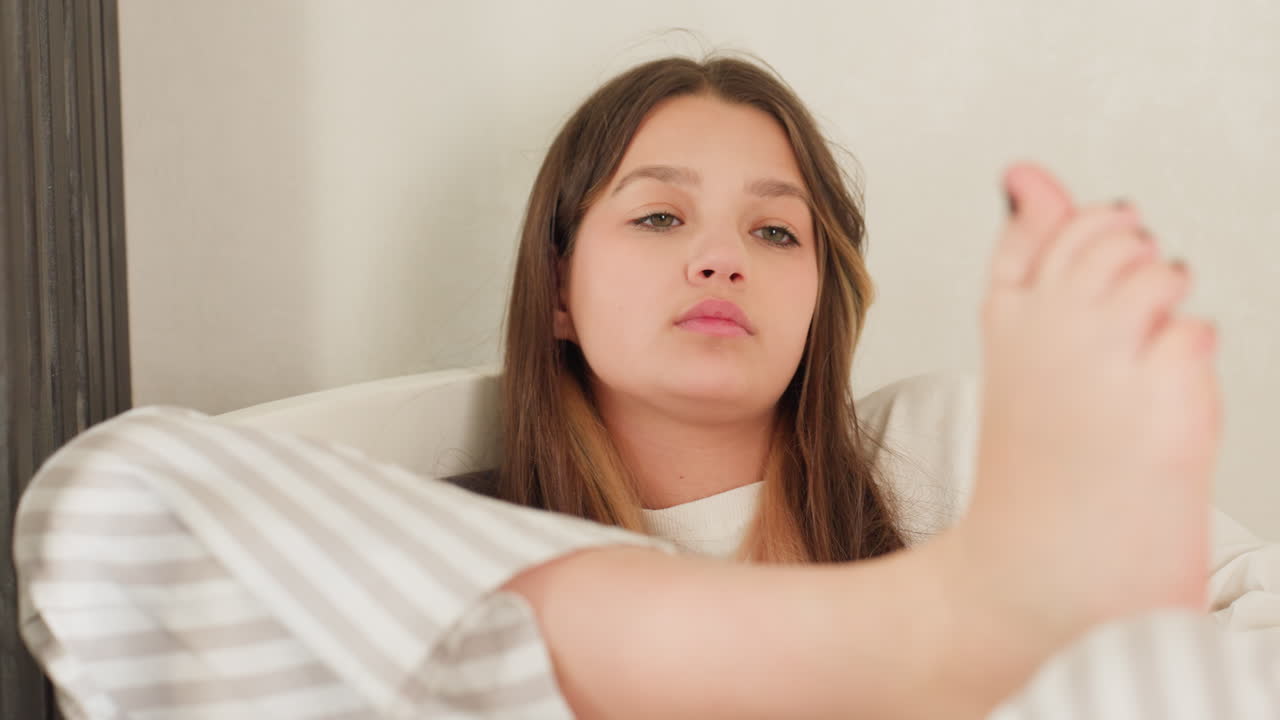 Elegant young girl in striped pajamas sits with legs crossed, gazing thoughtfully at raised leg, calm bedroom setting, soft daylight on face, serene mood, intimate closeup capturing quiet self care