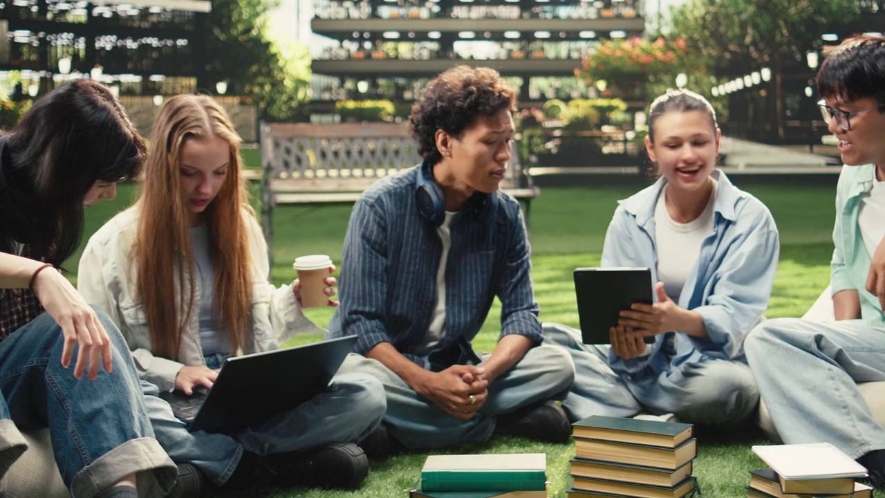 Diverse students checking notes on tablet and laptop with stack of books