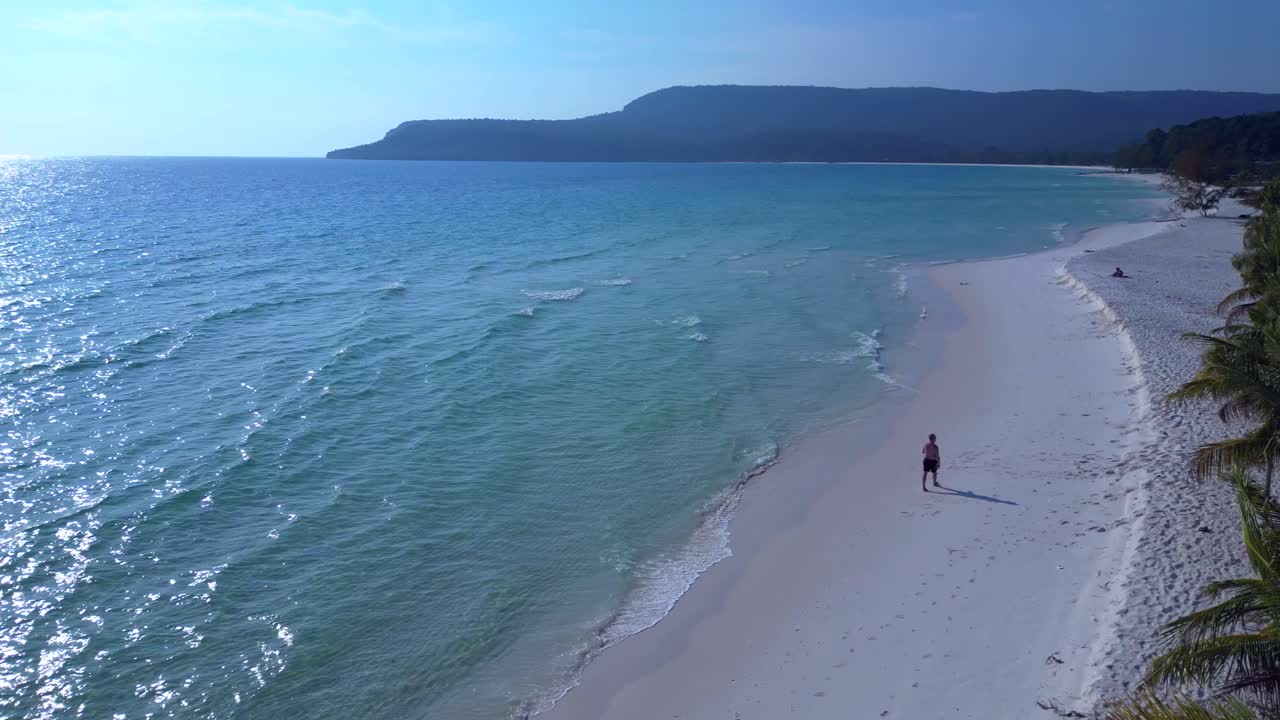 Crystal clear turquoise water and white sand beach, lined with lush palm trees and a lone tourist enjoying the serene tropical paradise. Fabulous aerial view flight descending drone
