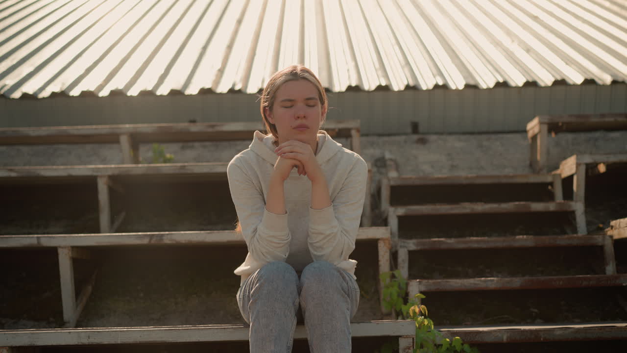 Thoughtful woman in casual attire sits on rustic stadium bleachers, hands joined together, with a pensive expression, looking contemplative in the soft outdoor light