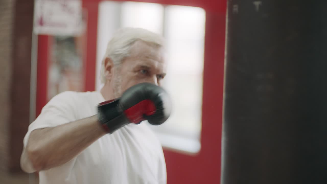 Uomo dai capelli grigi che si allena con calci al sacco da boxe in un club di arti marziali. Concetto di anziano attivo.
