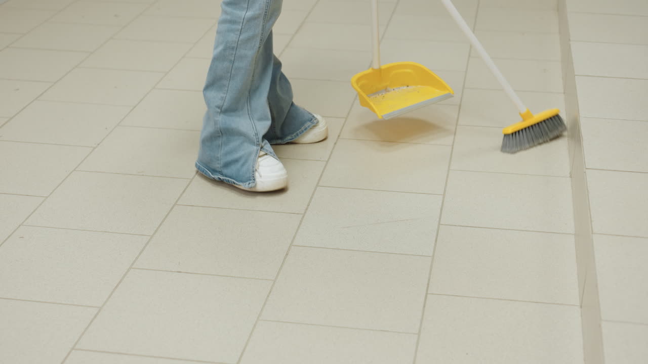 Leg view shows laundry worker in white sneakers and jeans sweeping tiled floor with broom and dustpan beside industrial washers, keeping clean interior, routine sanitation service, motion by step