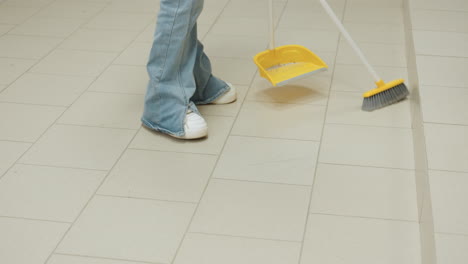 leg view shows laundry worker in white sneakers and jeans sweeping tiled floor with broom and dustpan beside industrial washers, keeping clean interior, routine sanitation service, motion by step