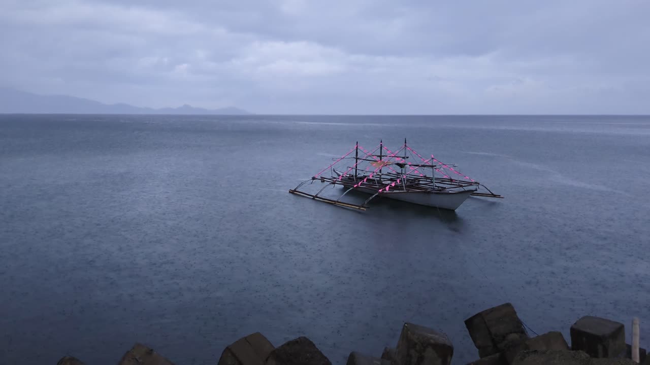 Static shot of lone, Philippine, traditional bangka boat anchored along the calm ocean coast during an overcast, rainy day.