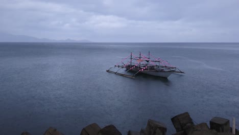 static shot of lone, philippine, traditional bangka boat anchored along the calm ocean coast during an overcast, rainy day.