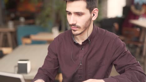 un joven apuesto con barba y bigotes está sentado en un café con auriculares, escribiendo en una laptop frente a él y sonriendo para sí mismo