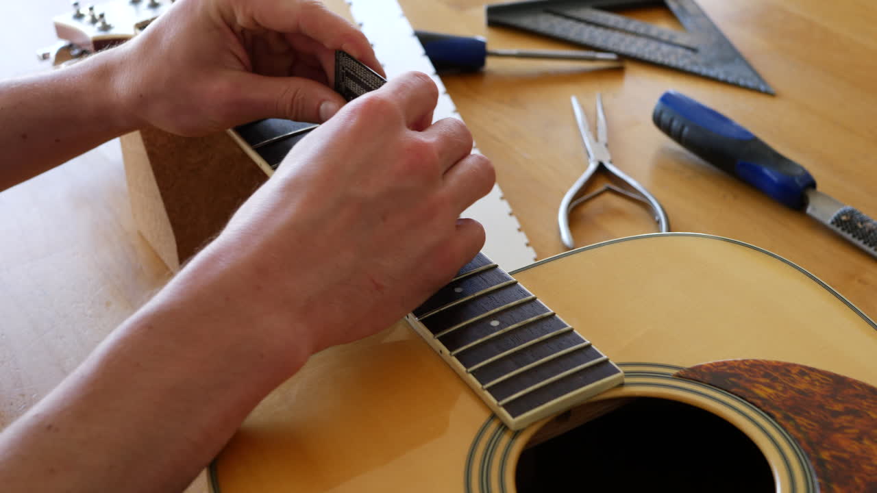 Hands of a luthier craftsman measuring and leveling an acoustic guitar neck fretboard on a wood workshop bench with lutherie tools