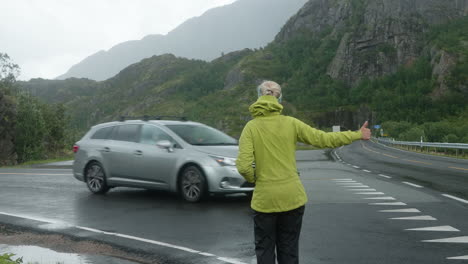 pretty blonde girl hitchhiking at the side of the road in the lofoten islands on a cold cloudy rainy day, cars are passing, norway