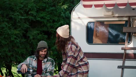 una chica morena feliz con una camisa a cuadros trae pasteles frescos para su novio moreno con una camiseta a cuadros verde. y el tipo vierte té caliente en una taza y abraza a su novia cerca del remolque en el campamento durante su picnic fuera de la ciudad en el verano