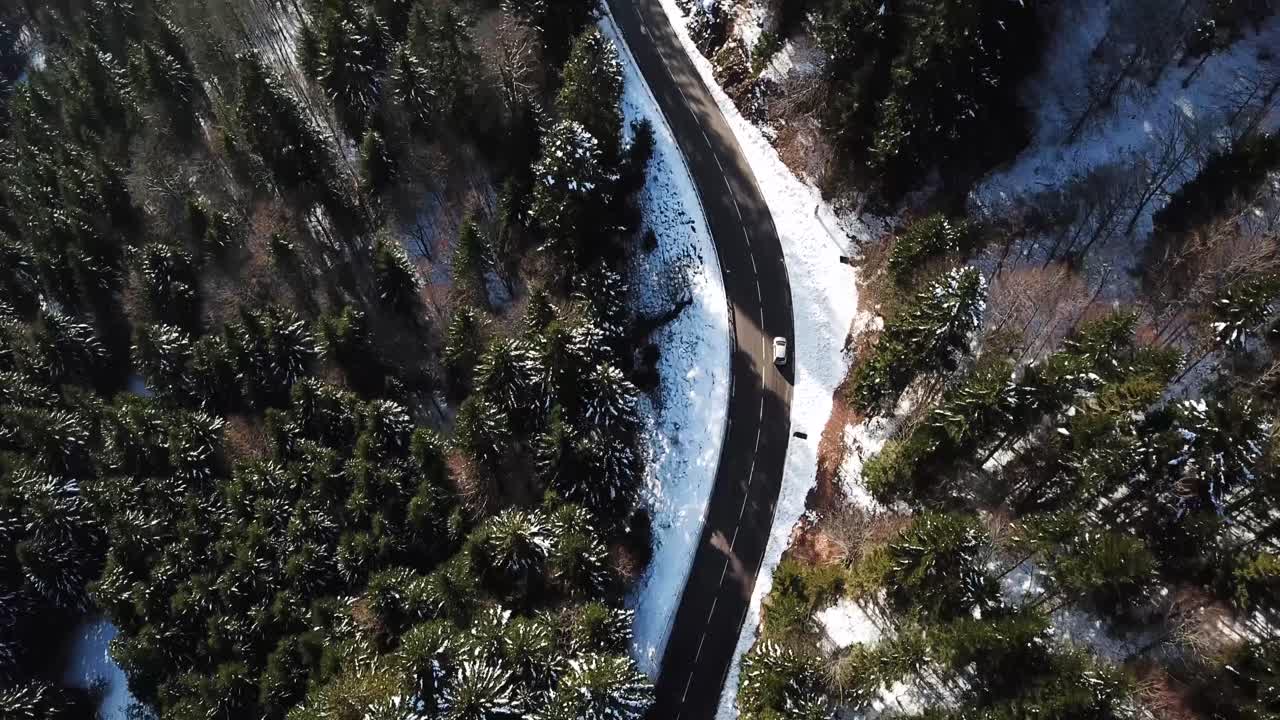 Aerial wide view of a car driving on a mountain road in a snowy winter landscape with green pine trees surrounding it