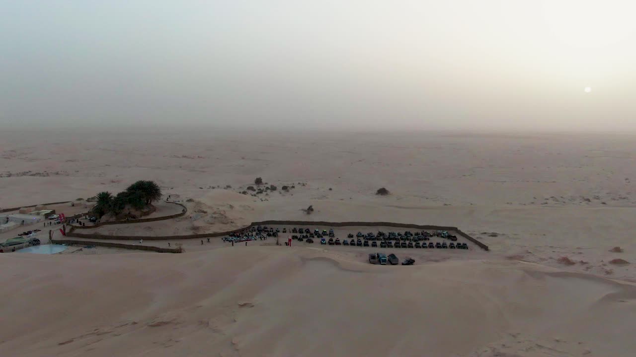 Vista aerea di un campo nel deserto organizzato con veicoli fuoristrada all'alba, che mostra vaste pianure sabbiose, una leggera foschia e un raduno strutturato in un ambiente arido e isolato.