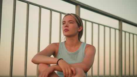 young lady sitting outdoors near iron railing at dusk, touching her hair with serene expression, wearing black bangle on wrist, arms crossed over knees while looking thoughtfully into distance