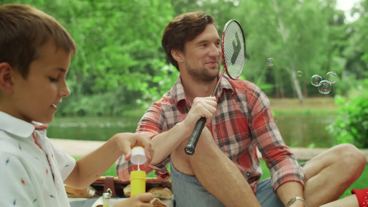 남자 와 여자 는 바 ⁇  에서 배드민턴 라켓 을 들고 있다. 아이 들 은 비누 거품 을 불고 있다