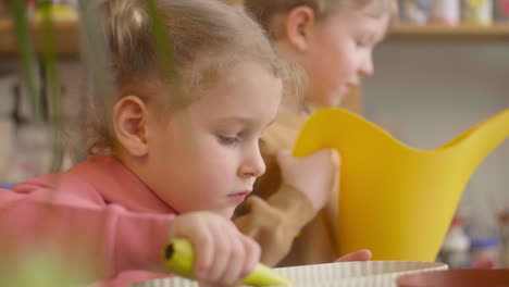 vista de cerca de una niña rubia y un niño rubio preparando el suelo para regar una olla sentada en una mesa donde hay plantas en un taller de artesanía