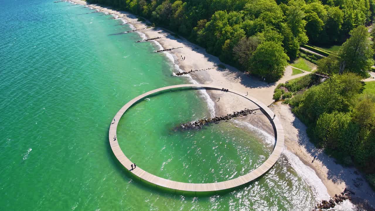Aerial top-down view of a circular pier structure stretching into shallow emerald waters, bordered by sandy shoreline and lush green trees