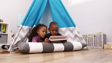 two children lying in tent in playroom with digital tablet