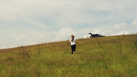 long haired blonde girl running up a hill on grass in slow motion with clouds in the background, drone follow cam