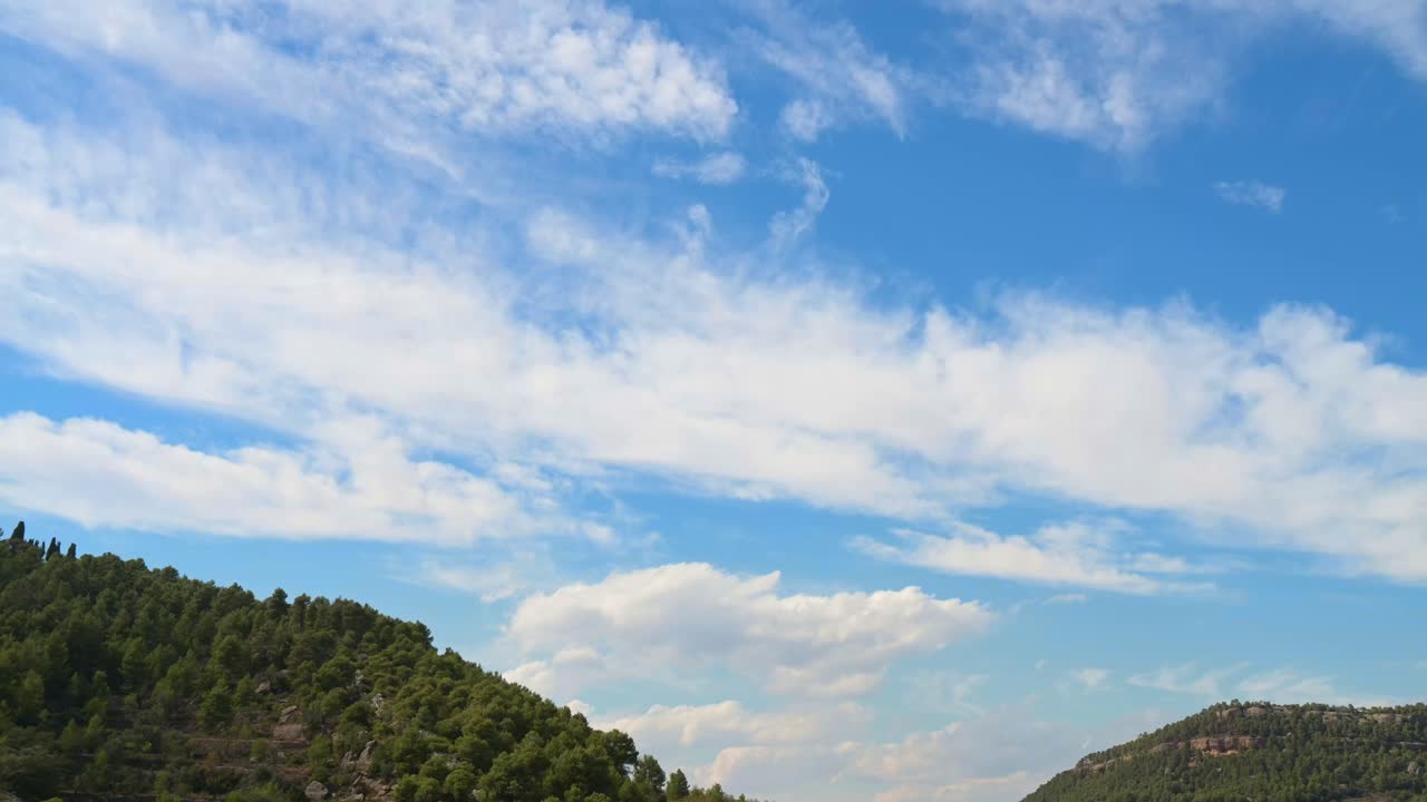 Delicate wispy cirrus time lapse clouds float elegantly across a vivid blue sky, high above a peaceful mountain valley nestled among lush green pine trees