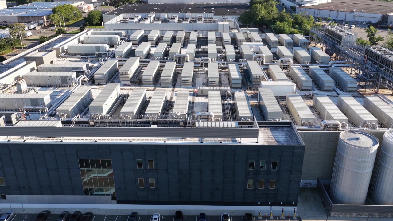Filas de unidades de refrigeración industrial cubren el tejado de un centro de datos en Secaucus, Nueva Jersey; una fachada de metal oscuro se eleva junto a un alto tanque de almacenamiento plateado y edificios de almacén distantes. Vista aérea amplia de dron desde arriba.