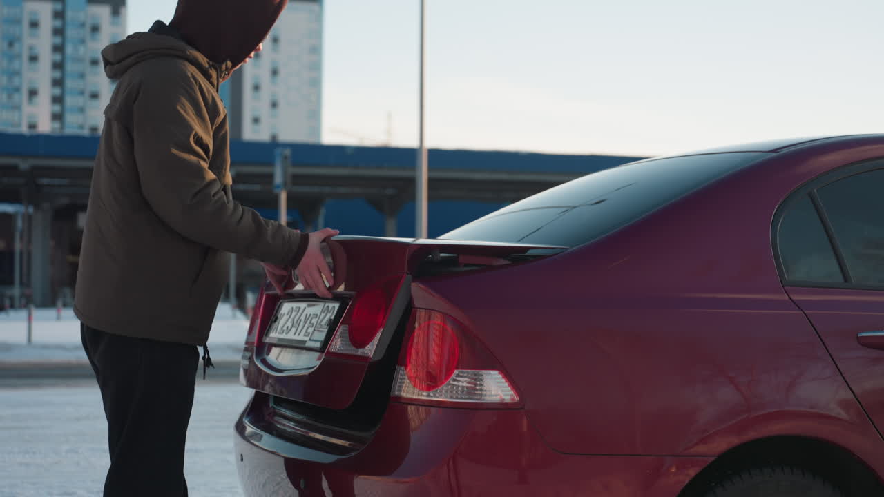 Young adult in hooded jacket closes car boot with one hand and turns to look at camera in snowy parking area with city buildings and vehicles in background under daylight