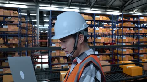 close up side view of asian male engineer with safety helmet working on a laptop while standing in the warehouse with shelves full of delivery goods